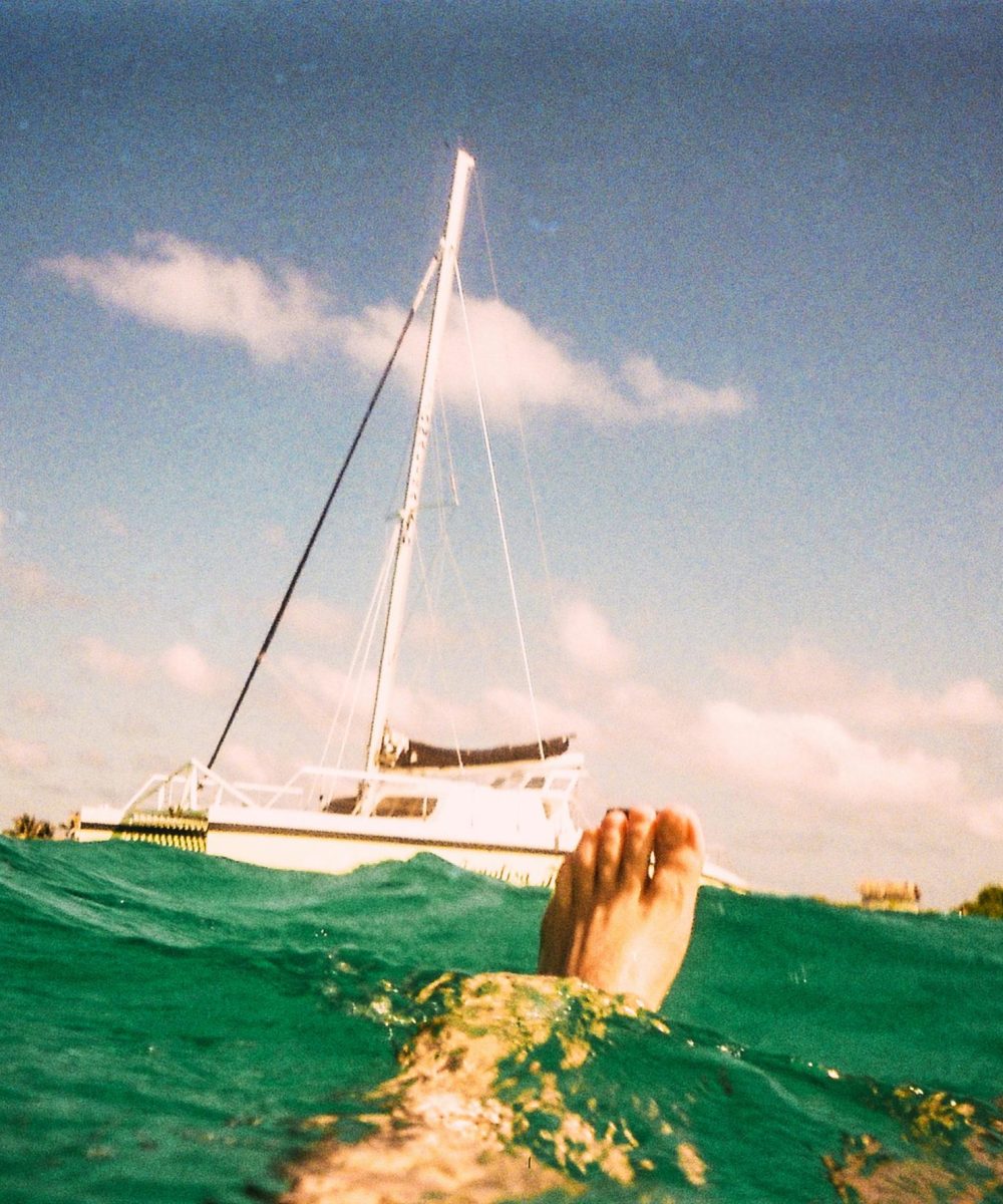 A person floating in turquoise water near a sailboat under a sunny sky.