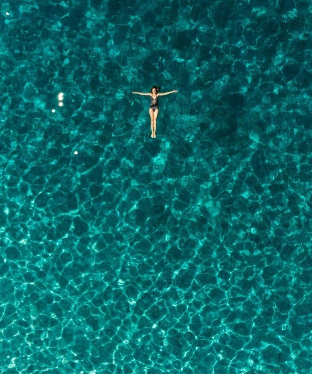 Drone shot of a woman floating in clear blue waters next to a boat in Greece.
