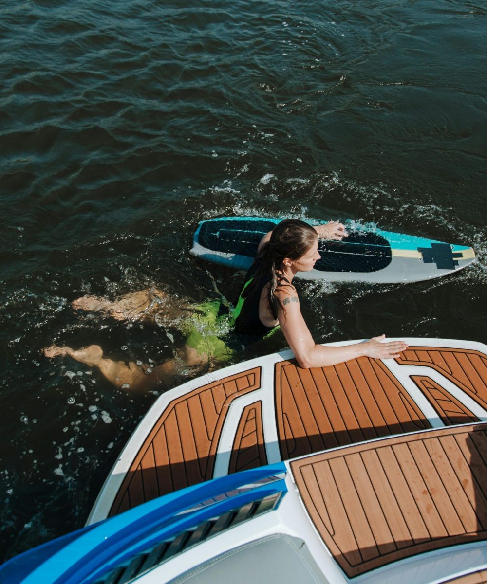 A woman with surfboard in water beside a boat on a sunny summer day.